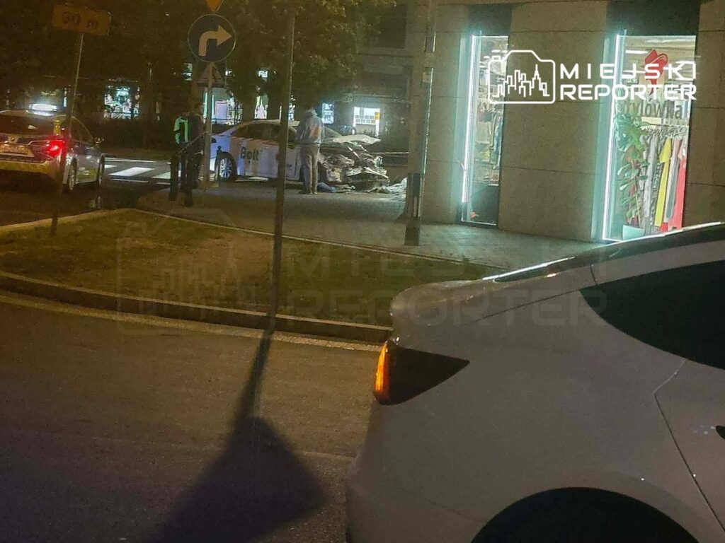 Policjant rozmawia z kierowcą samochodu osobowego na skrzyżowaniu, w tle widoczne sklepy z oświetleniem neonowym.