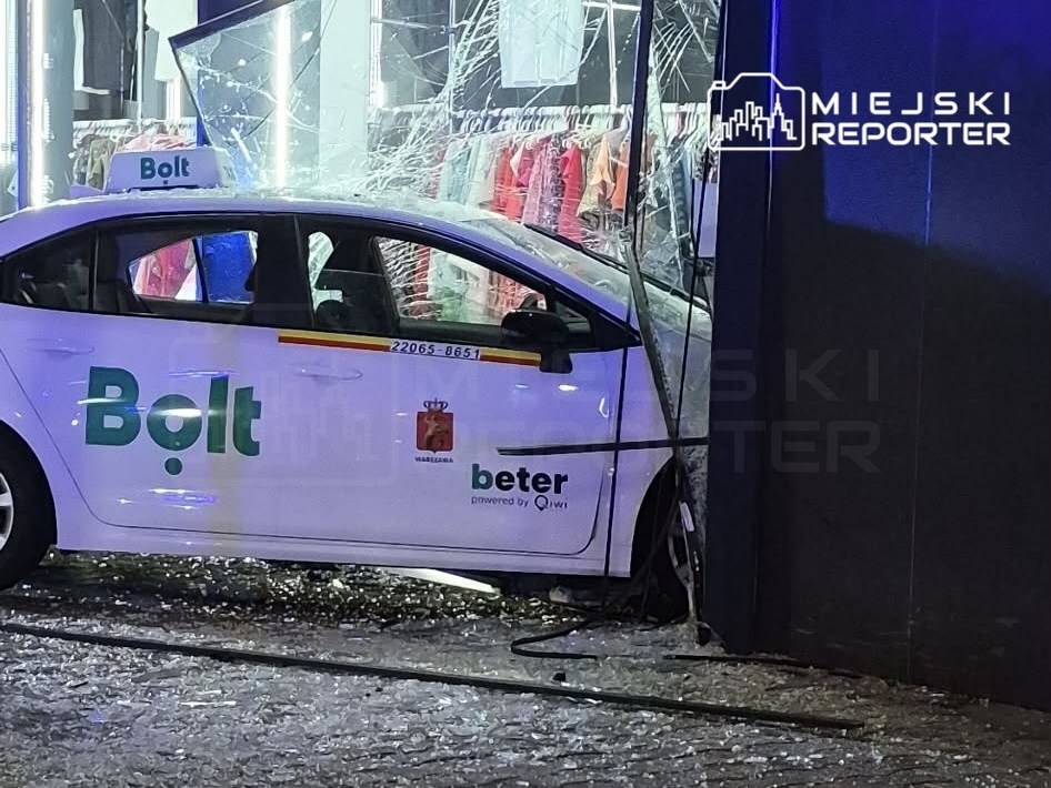 Zniszczony samochód osobowy marki Bolt wjechał w witrynę sklepową, a szkło leży na chodniku.