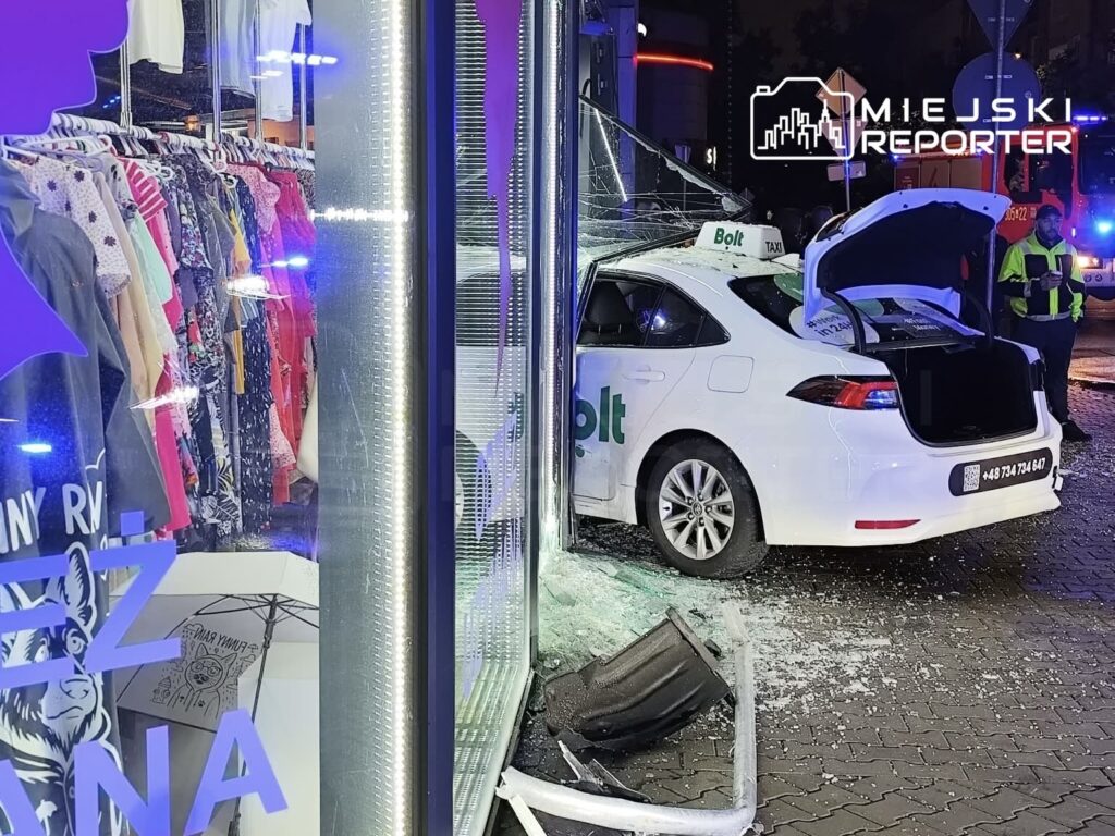 Biały samochód taksówki Bolt uderzył w witrynę sklepu, a szyby są rozbite, wokół leżą fragmenty szkła.