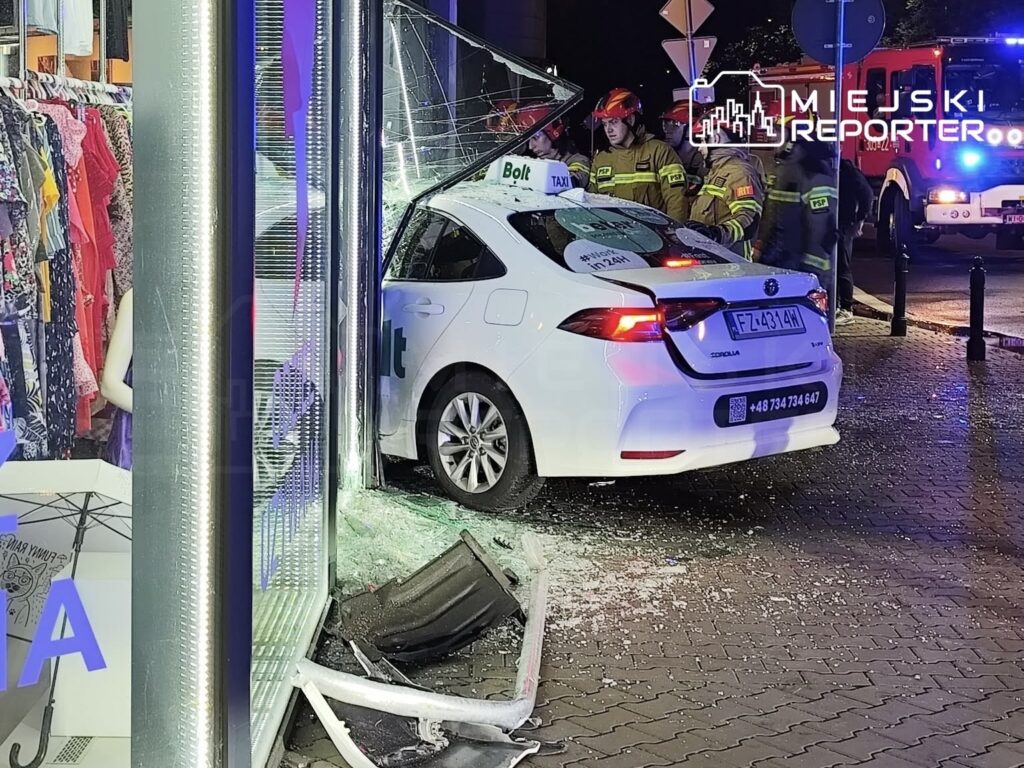 Biały samochód osobowy wjeżdża w witrynę sklepową, wokół rozbite szkło i zniszczone elementy. Strażacy udzielają pomocy.