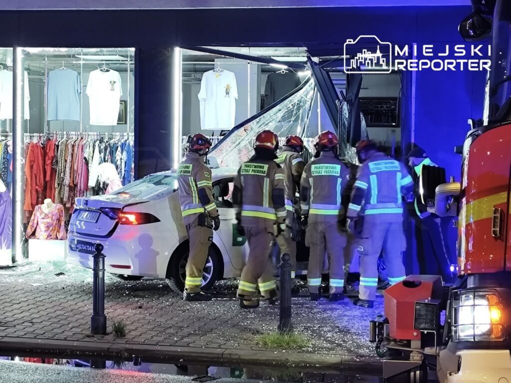 Grupa strażaków w mundurach przeszukuje wnętrze sklepu odzieżowego po wypadku samochodowym, w którym pojazd uderzył w witrynę.