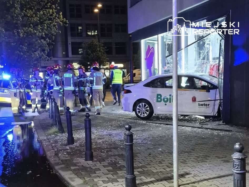 Zespół strażaków oraz policjantów pracuje przy uszkodzonym budynku, w który wjechał samochód.