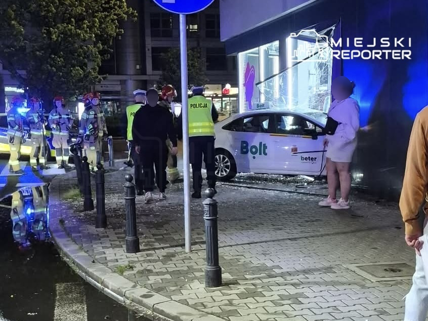 Samochód Bolt wjeżdża w witrynę sklepu, obok grupka strażaków i policjantów w akcji.