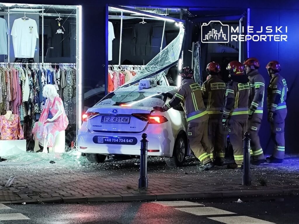 Samochód osobowy wjechał w witrynę sklepową, a obok niego stoi pięciu strażaków w mundurach, oceniających sytuację.