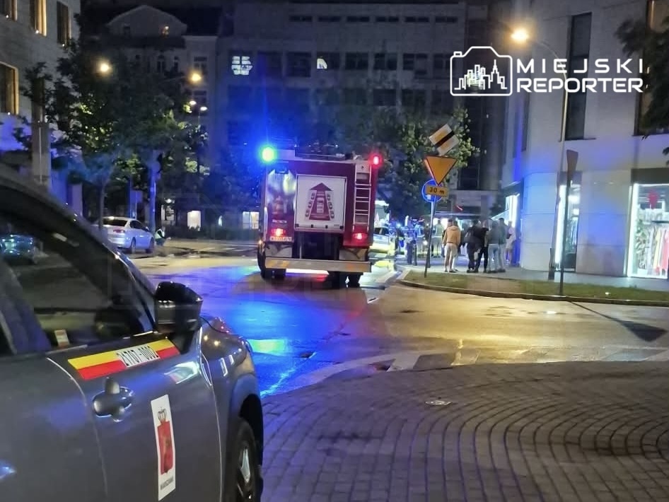 W nocy, w mieście, straż pożarna z włączonymi sygnałami świetlnymi przejeżdża obok grupy ludzi na chodniku.