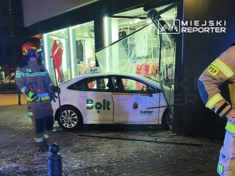 Samochód marki Bolt wjechał w witrynę sklepu, a obok stoją strażacy w pełnym umundurowaniu.