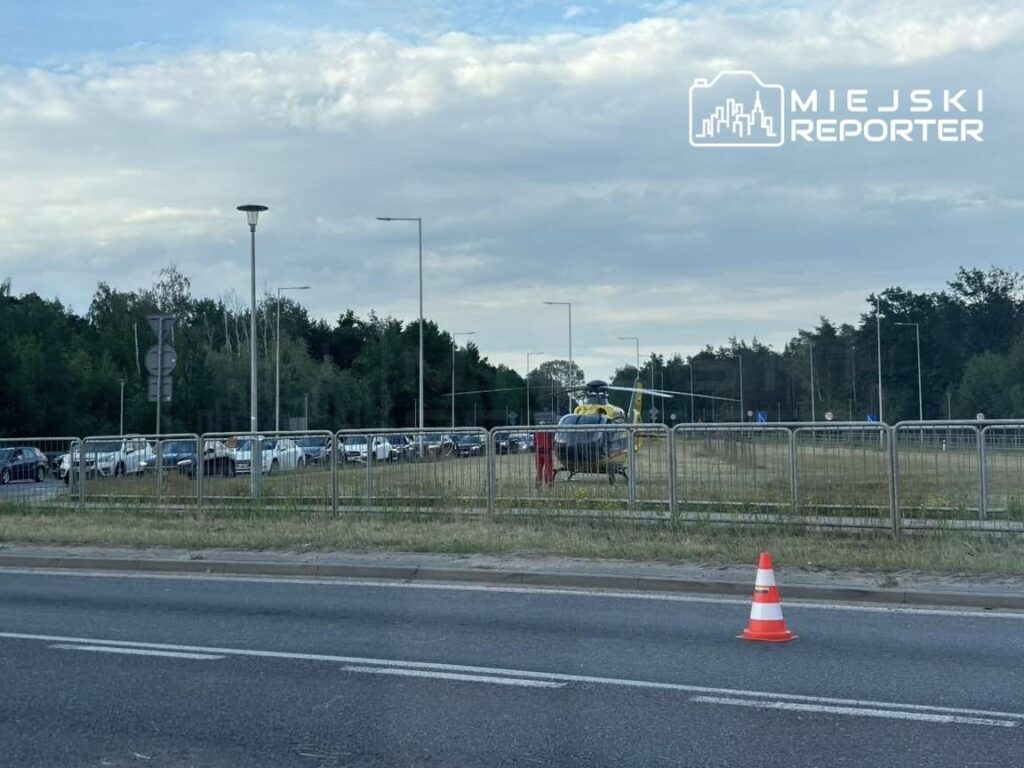 Helikopter lądował na trawniku obok drogi, w tle widoczne samochody stojące w korku.