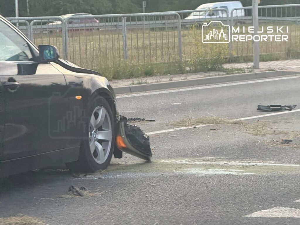 Czarny samochód osobowy z uszkodzoną oponą stoi na drodze, wokół leżą fragmenty pojazdu i trawa.