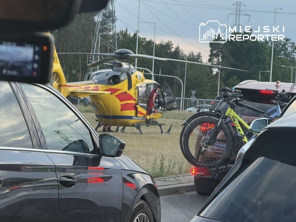 Lądowisko helikoptera ratunkowego obok zablokowanej drogi, w tle widoczne samochody i rowery na przyczepce.