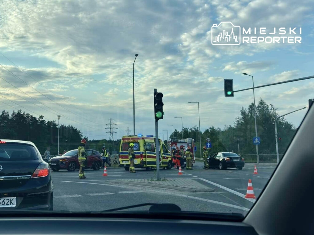 Karetka i strażacy udzielają pomocy przy zderzeniu dwóch samochodów na skrzyżowaniu, w tle sygnalizacja świetlna.