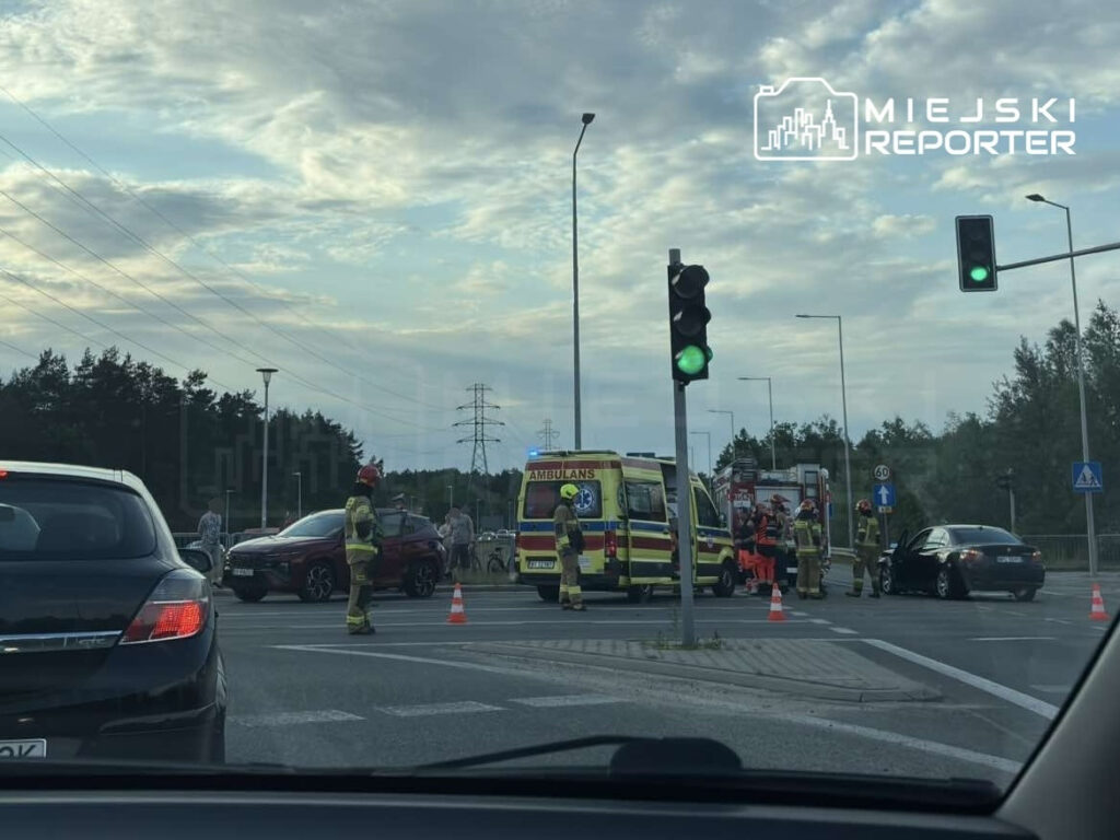 Grupa strażaków oraz ratowników medycznych pracuje przy wypadku drogowym, na drodze z sygnalizacją świetlną.