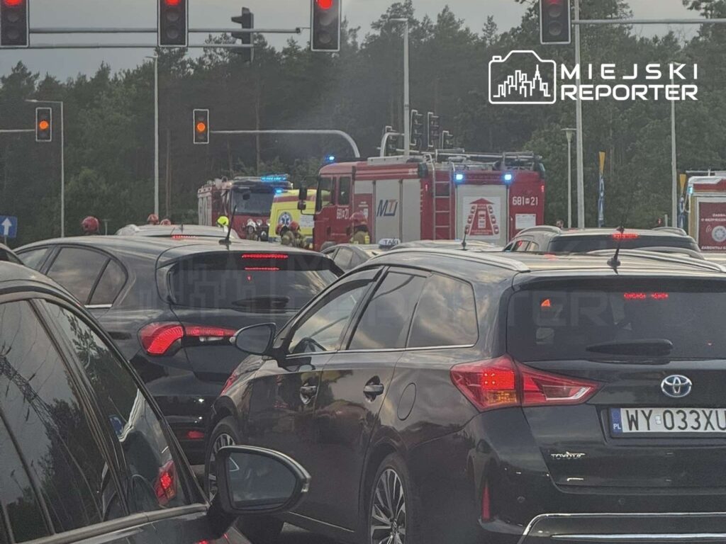 Ruch uliczny zatrzymany na skrzyżowaniu, w tle widać wozy strażackie z włączonymi sygnałami świetlnymi.