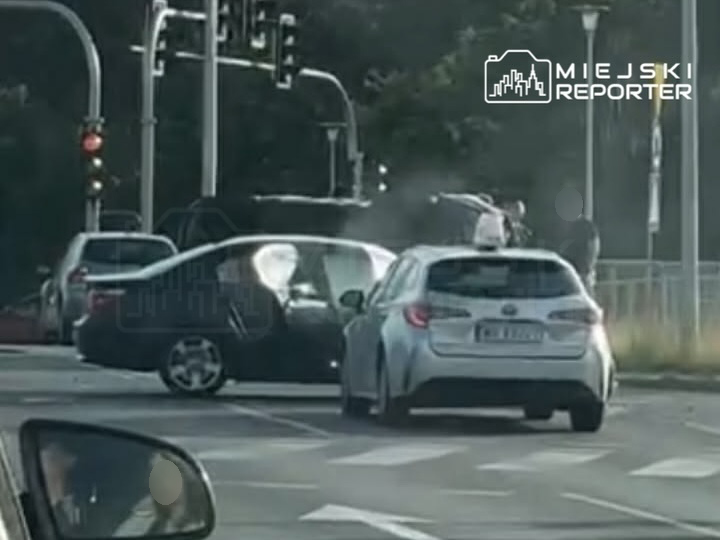 Dwa uszkodzone samochody na skrzyżowaniu, jeden z otwartymi drzwiami, w tle sygnalizacja świetlna.