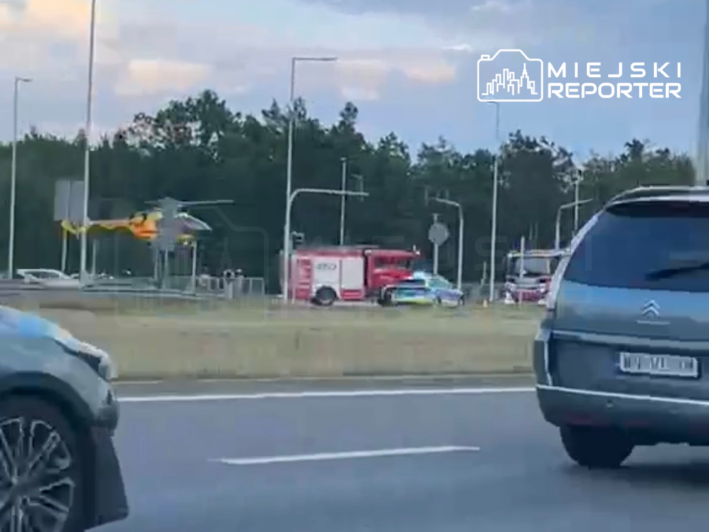 Helikopter ratunkowy ląduje obok karetki i radiowozu na skrzyżowaniu przy drodze. W tle widać inne pojazdy.