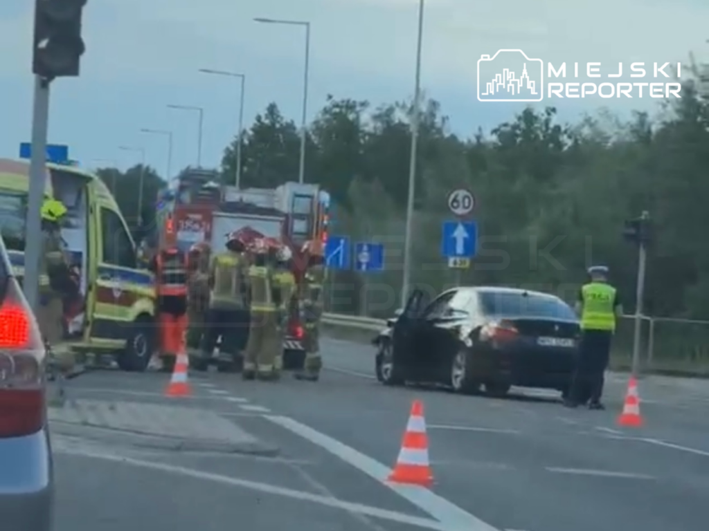 Na drodze stoją strażacy i policjant, obok uszkodzony samochód, w tle karetka i woz strażacki.