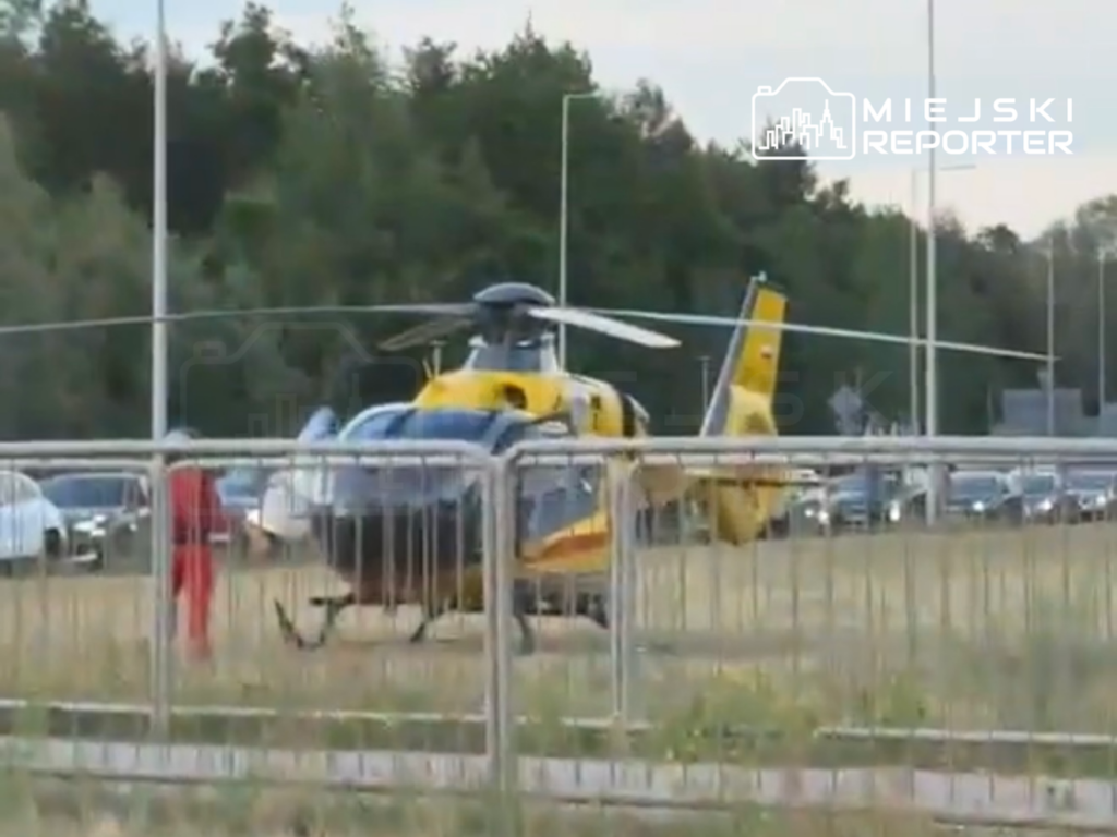 Helikopter ratunkowy lądował na trawniku obok ruchliwej drogi, a ratownik w czerwonym stroju przygotowuje się do akcji.