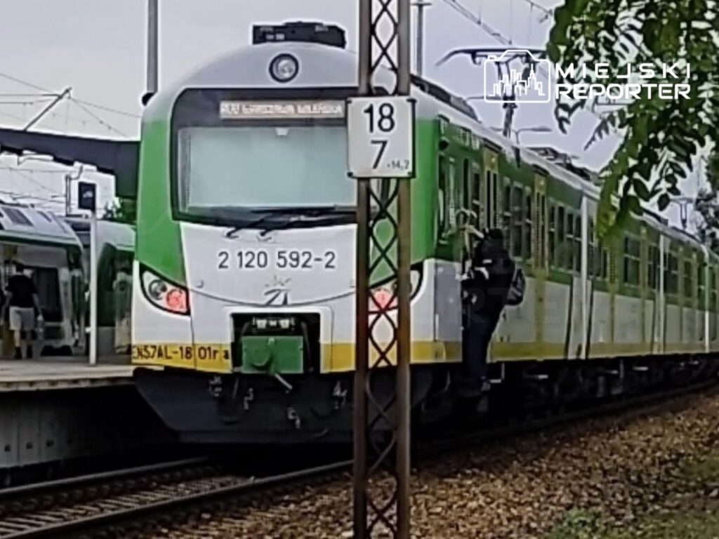 Pasażer wsiada do zielonego pociągu na stacji kolejowej, obok widoczny drugi pociąg.