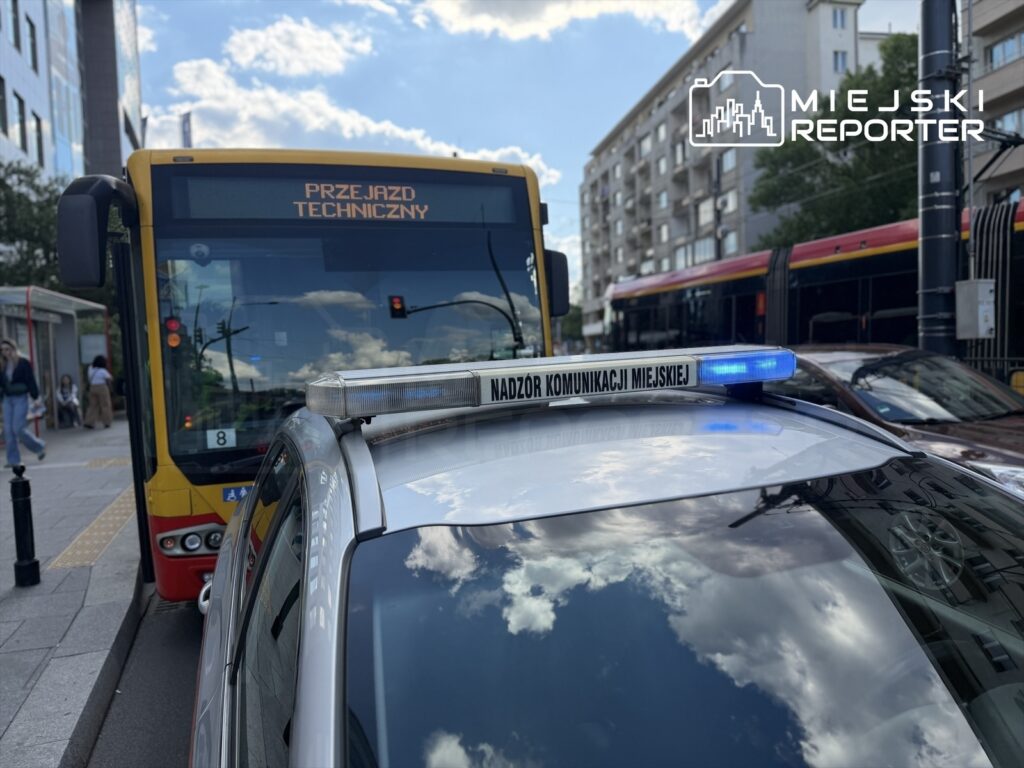 Policyjny radiowóz z niebieskim światłem stoi obok autobusu miejskiego z napisem "PRZEJAZD TECHNICZNY" w tle.