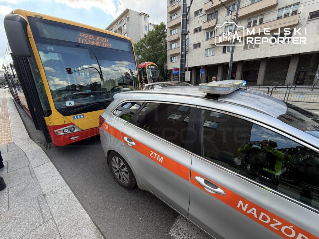 Autobus przegubowy zatrzymany na ulicy, obok policyjny samochód z włączonymi sygnałami świetlnymi.