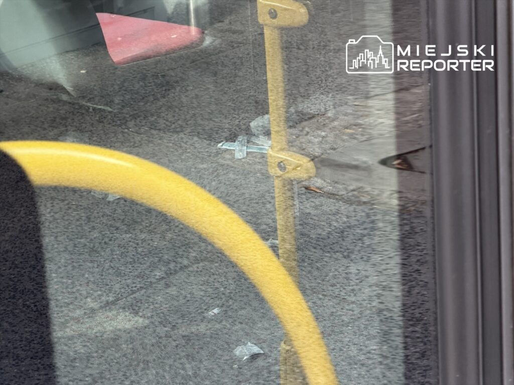 Żółty uchwyt na poręczy autobusu widoczny przez szklane drzwi, w tle widać czerwone siedzenie.