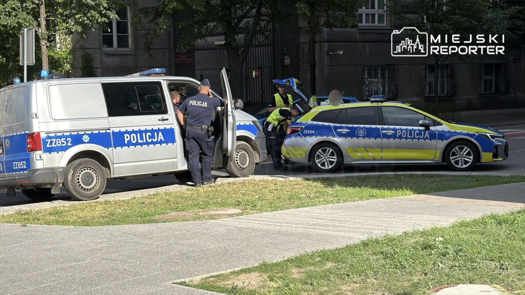 Policjanci w mundurach przeszukują dwa radiowozy na ulicy, obok jednego z pojazdów stoi otwarta drzwi.