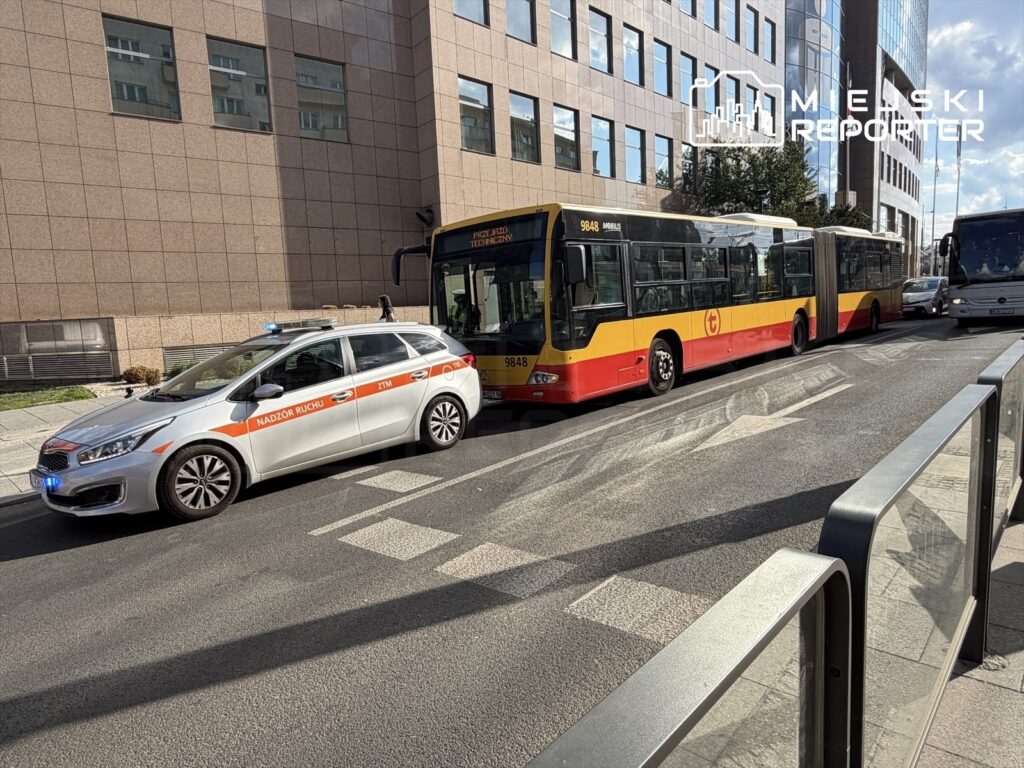 Radiowóz z niebieskimi światłami stoi obok przegubowego autobusu w miejskim otoczeniu.