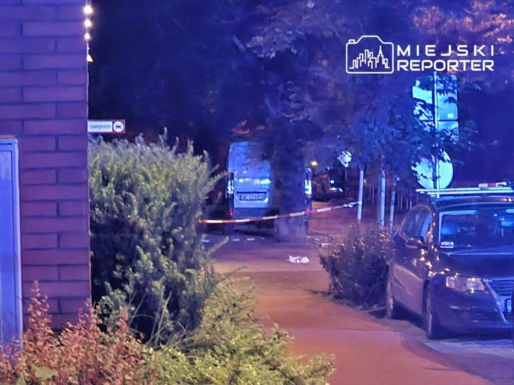 Na chodniku w nocy widoczny jest radiowóz oraz taśma policyjna, otaczająca miejsce zdarzenia.