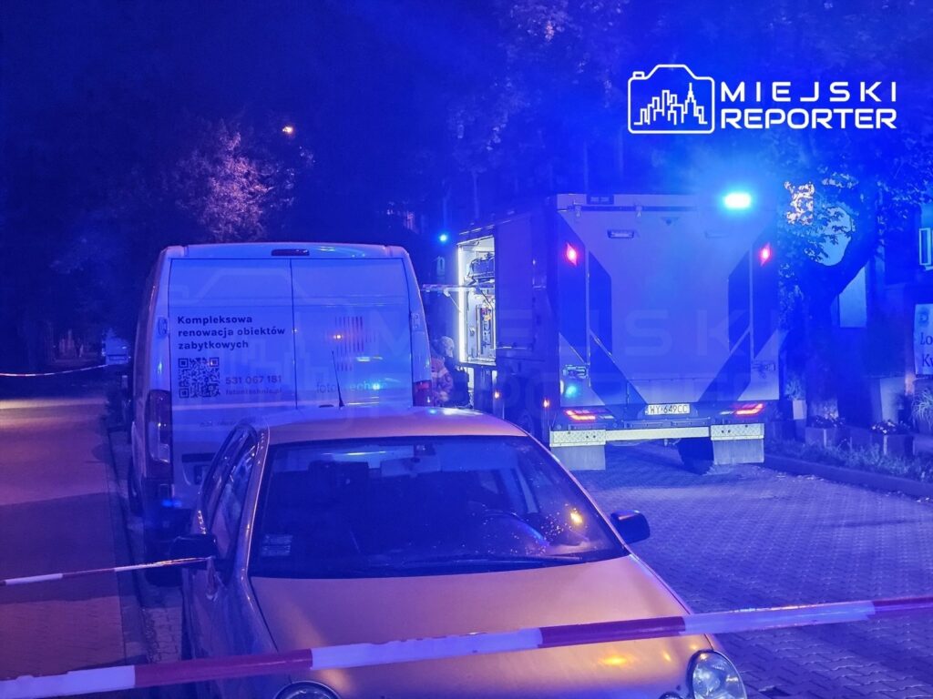 Na ulicy stoją dwa pojazdy: biały van z napisem "Kompleksowa renowacja obiektów zabytkowych" oraz nieoznakowany pojazd służbowy z włączonymi światłami. W tle widać oświetlone drzewa.