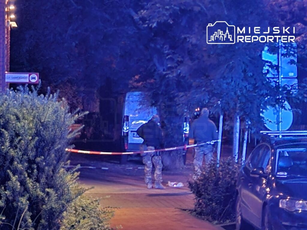 Dwaj funkcjonariusze w mundurach stoją przy taśmie zabezpieczającej w oświetlonym niebieskim świetle, obok zaparkowanego pojazdu.