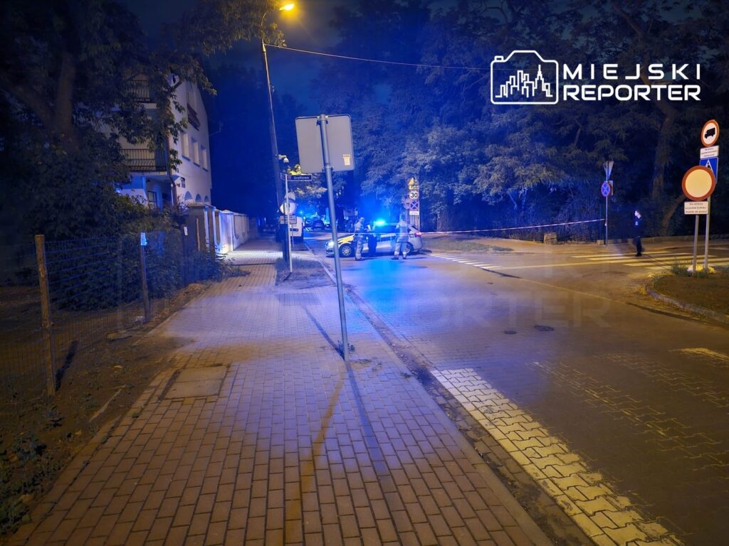 Nocna scena na ulicy z policyjnymi radiowozami i niebieskimi światłami, zamknięty odcinek drogi.