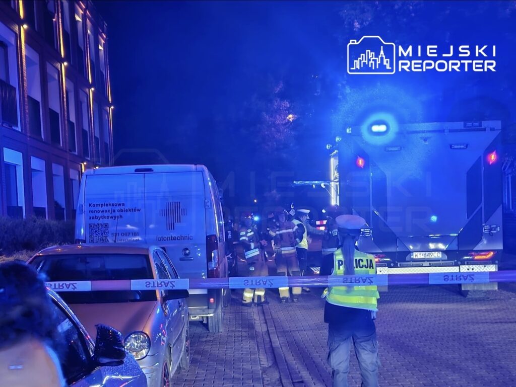 Grupa strażaków i policjantów pracuje na miejscu zdarzenia, obok zaparkowanych pojazdów, w oświetlonym niebieskim świetle.