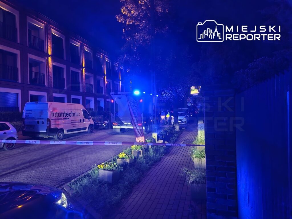 Niebieskie światła policyjne oświetlają ulicę, gdzie zaparkowane są pojazdy służbowe oraz oznakowane miejsce zdarzenia.