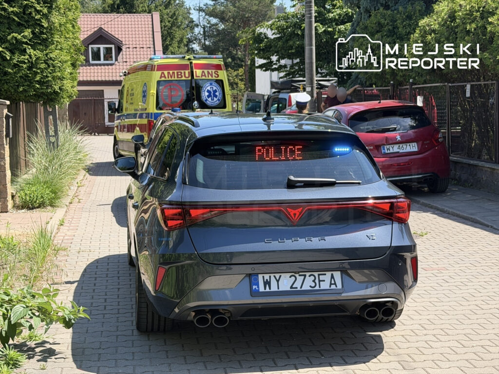 Czarny samochód z napisem "POLICE" na dachu stoi na wąskiej ulicy, obok karetki pogotowia.