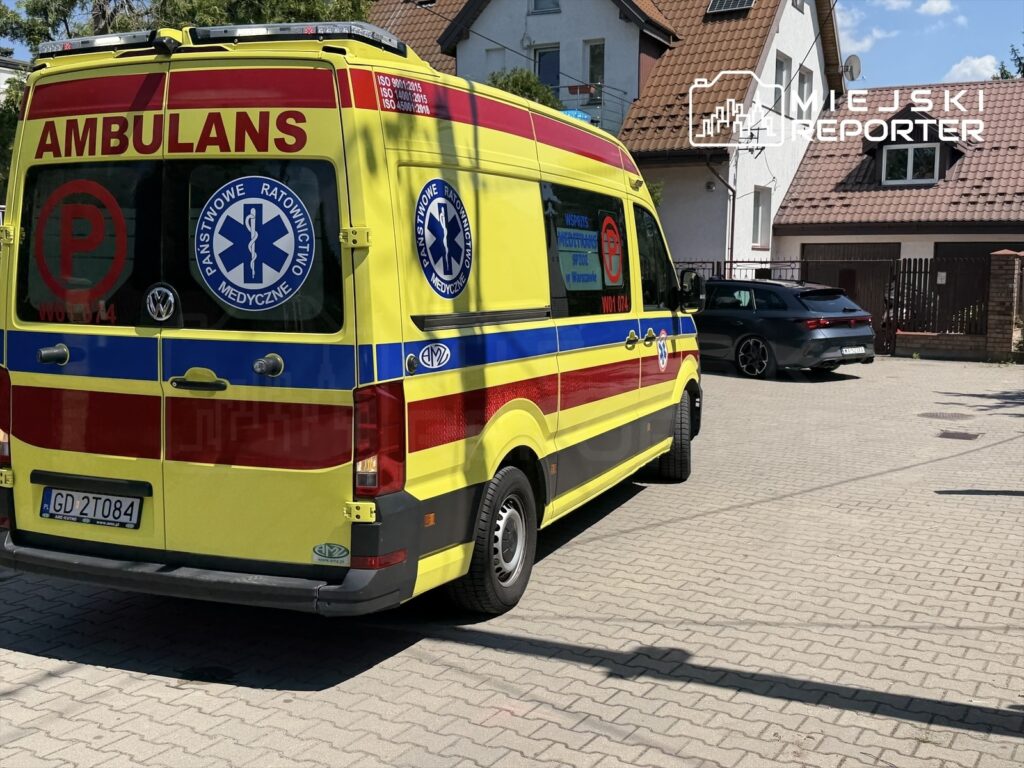 Karetka pogotowia z oznaczeniami "Panstwowe Ratownictwo Medyczne" zaparkowana na placu przed budynkiem mieszkalnym.
