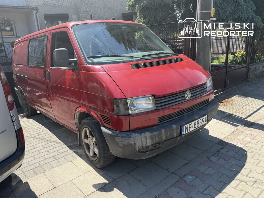 Czerwony Volkswagen Transporter zaparkowany na chodniku w słoneczny dzień, obok innych pojazdów.