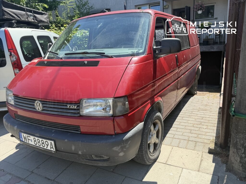 Czerwony furgon marki Volkswagen TDI zaparkowany na brukowanej nawierzchni obok budynku mieszkalnego.