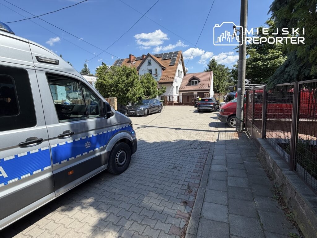 Policyjny van zaparkowany na wybrukowanej ulicy, w tle domy jednorodzinne i kilka zaparkowanych samochodów.