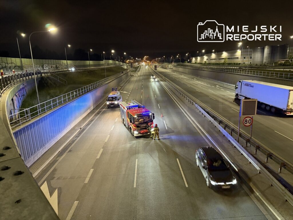 Strażacy pracujący przy wozie strażackim na nocnej autostradzie, obok zaparkowanego pojazdu i oznaczeń drogowych.