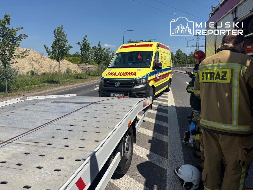 Ambulans z napisem "POMOC" zatrzymany przy drodze, obok strażacy w mundurach, przygotowujący się do akcji.