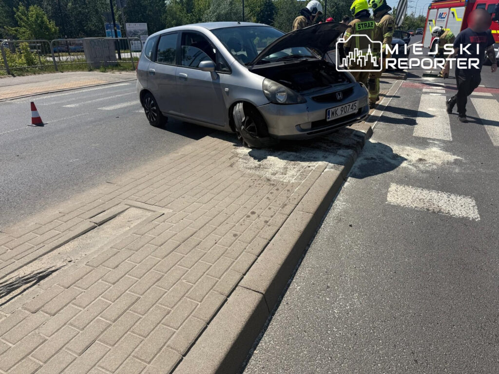 Samochód osobowy utknął na krawężniku, obok interweniują strażacy w odblaskowych strojach.