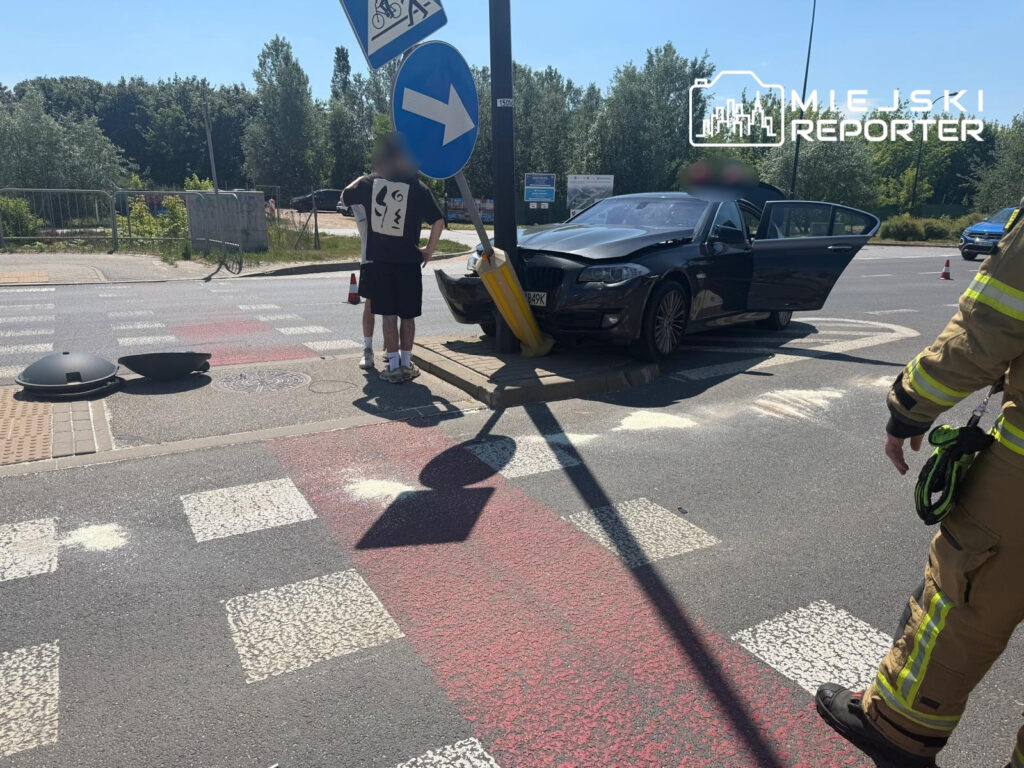 Mężczyzna w krótkich spodniach stoi przy uszkodzonym samochodzie, obok znaku drogowym oraz leżącym na ziemi elementem infrastruktury.