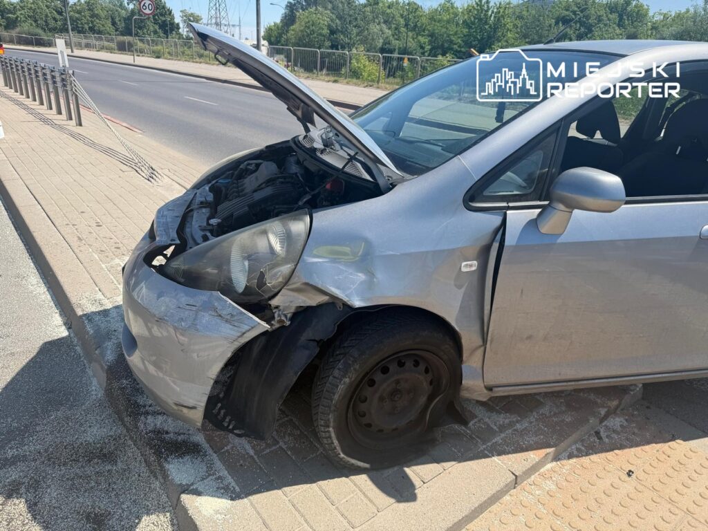 Uszkodzony srebrny samochód osobowy z otwartą maską stoi na chodniku obok drogi.