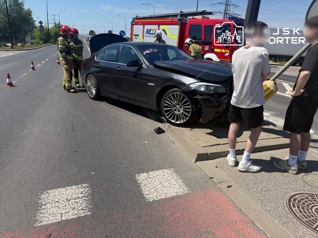 Czterech strażaków w mundurach omawia uszkodzone czarne auto na drodze, obok stoją dwaj mężczyźni.