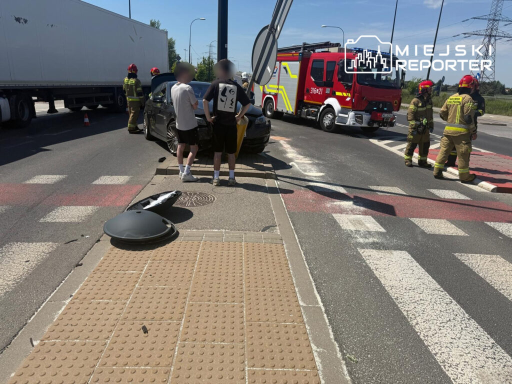 Dwaj mężczyźni stoją obok uszkodzonego samochodu, obok którego pracują strażacy i stoi wóz strażacki.