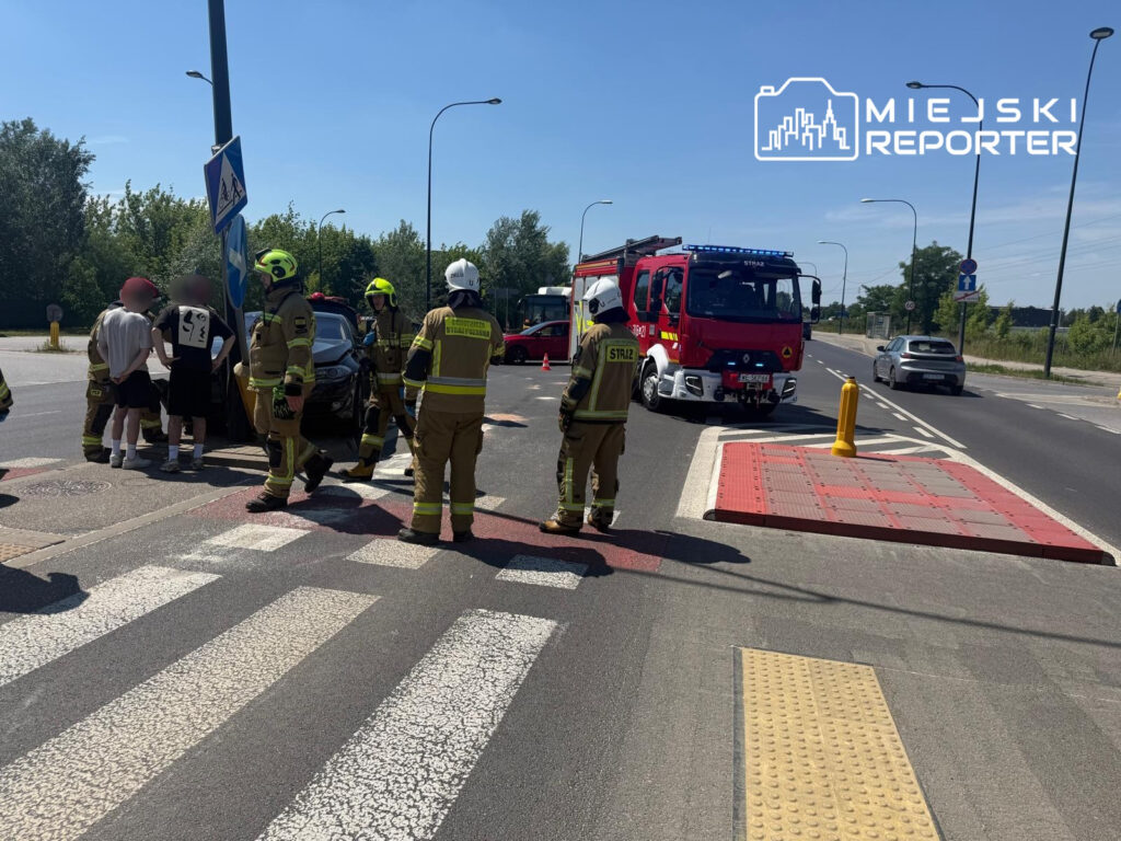 Grupa strażaków w mundurach omawia sytuację przy zderzeniu pojazdu na skrzyżowaniu, obok wozu strażackiego.