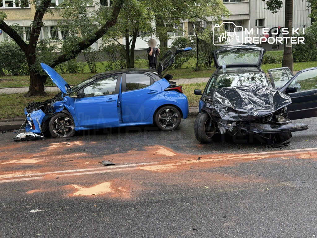 Niebieski hatchback z uszkodzoną przednią częścią stoi na jezdni, obok czarny samochód z zgniecionym przodem. Na asfalcie widoczne plamy płynów.