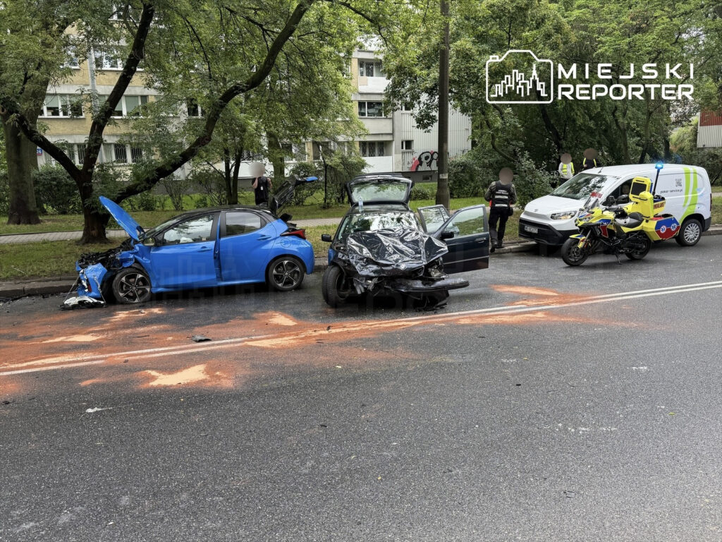 Dwa uszkodzone samochody na jezdni, w tym niebieski z otwartą maską, obok stoi pojazd służb ratunkowych.