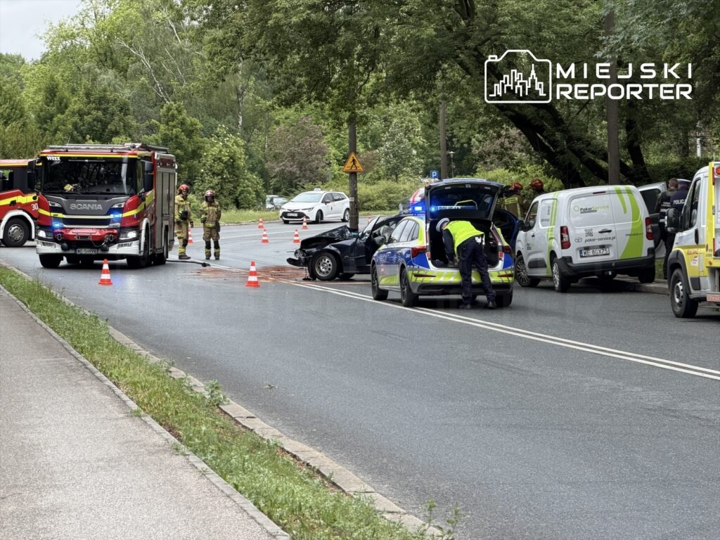 Na drodze zatrzymane pojazdy służb ratunkowych, w tym wóz strażacki i radiowóz, obok rozbite auto oraz kierowca sprawdzający bagażnik.
