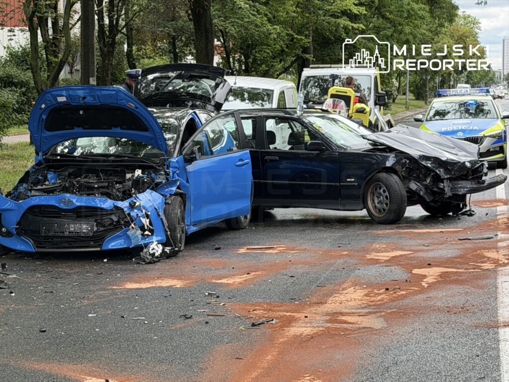 Dwa uszkodzone samochody po kolizji na jezdni, obok stoi policjant, a w tle widoczne pojazdy służb ratunkowych.