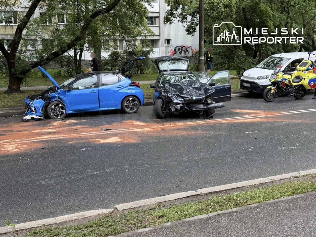 Zderzenie dwóch uszkodzonych samochodów na jezdni, obok stojących pojazdów służb ratunkowych.